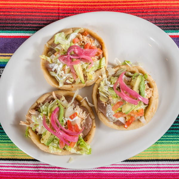 Three Chula on Broadway sopes with chicken, beans, lettuce, avocado, and pickled onions on a white plate.